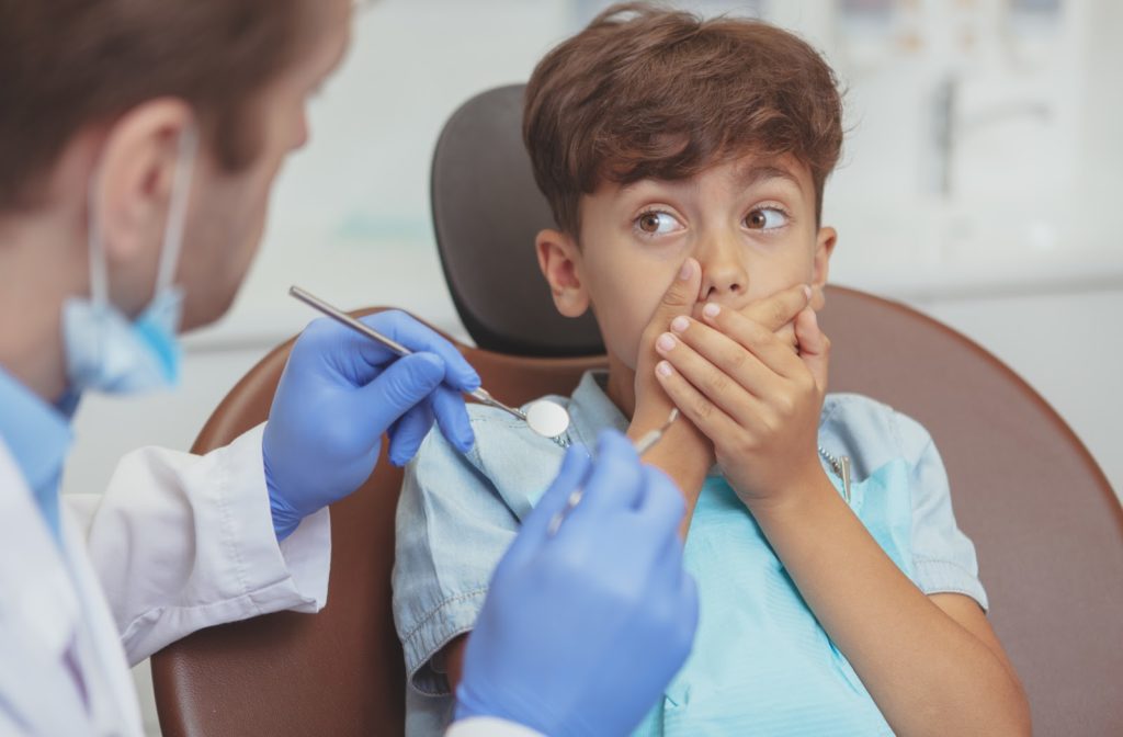 Wide-eyed boy covering mouth with both hands and staring at dentist during appointment
