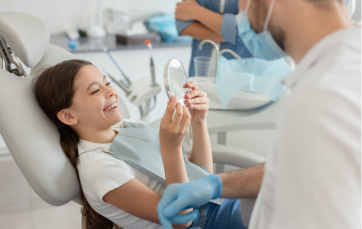 young girl holding mirror to look at her teeth after operation with her dentist present