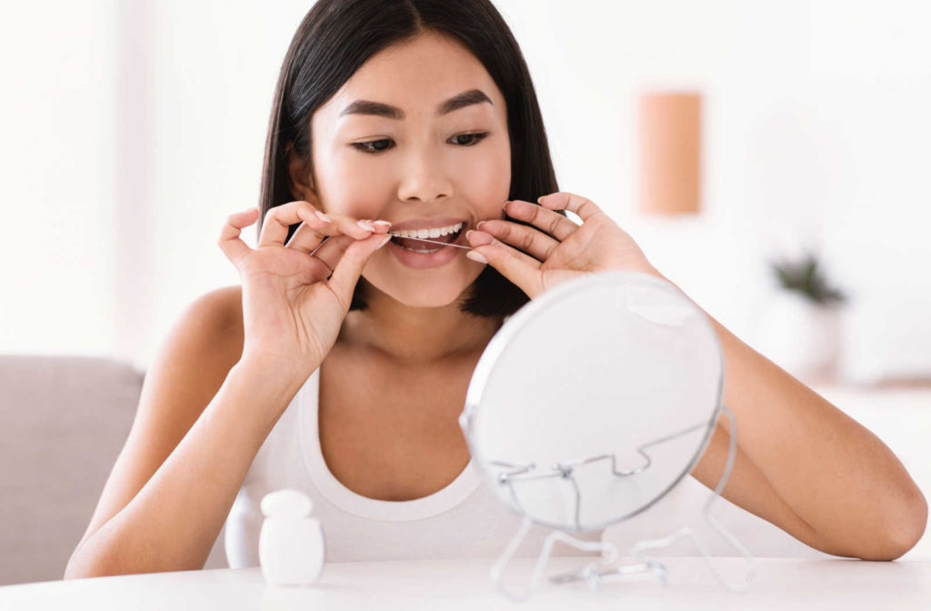 A woman looking in the mirror flossing her teeth around her retainer to maintain good oral hygiene.