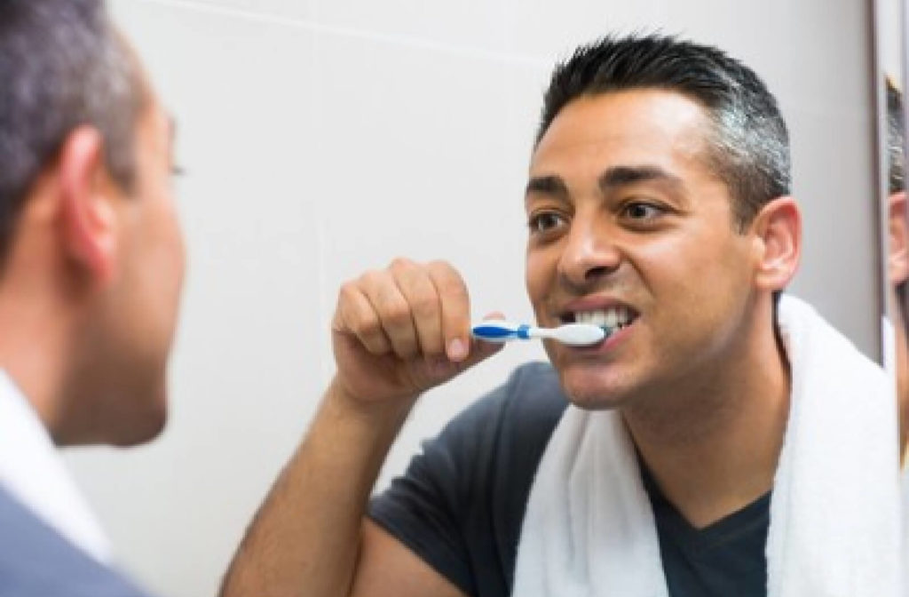 A man in the mirror in a blue shirt and a white towel on her neck is carefully brushing his teeth away from the tooth that was extracted.