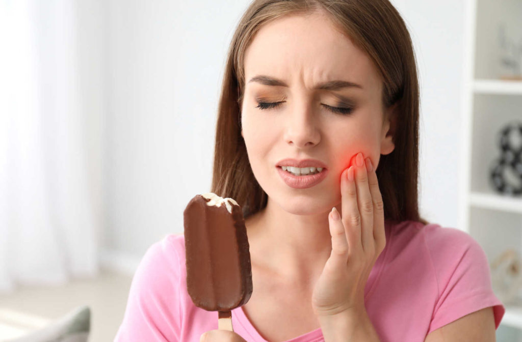 A woman holding an icecream bar in one hand and her cheek with the other as she experiences toothache due to sensitivity.