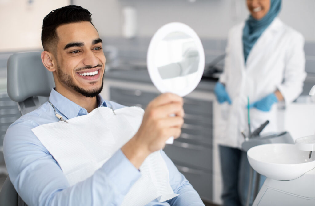 A man sitting in a dentist's chair, holding up a mirror and smiling with his dentist in the background.