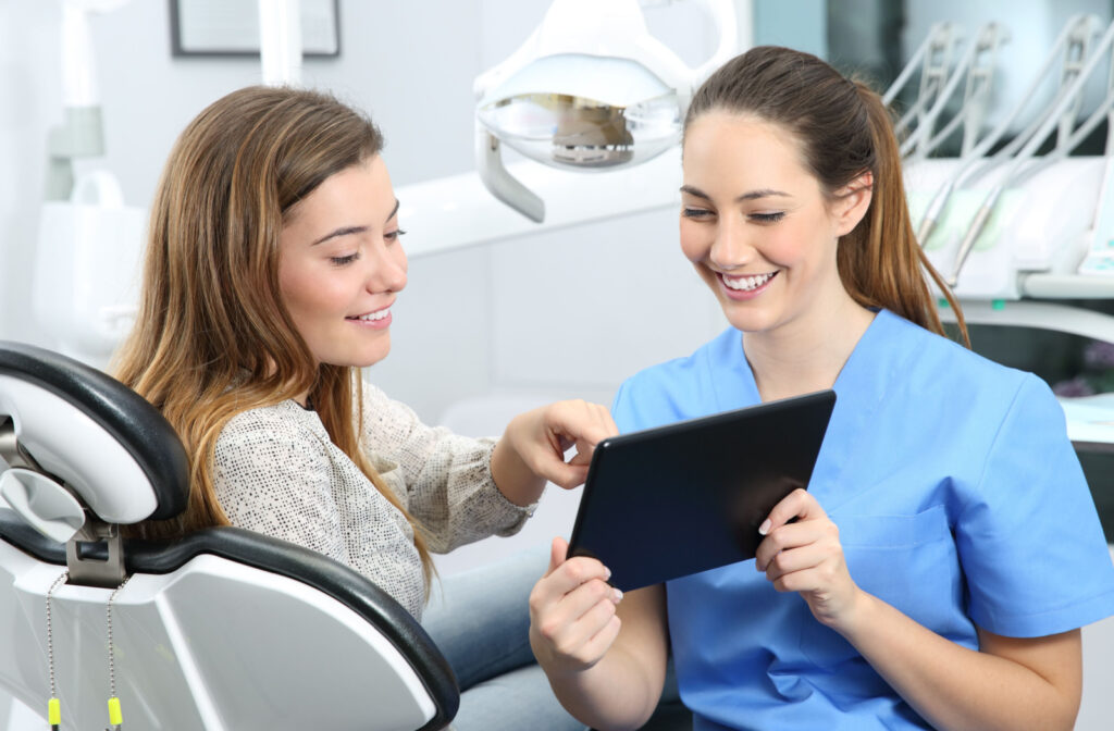 A young woman sitting in a dental chair and pointing to something displayed on a tablet as her dentist holds the tablet and smiles.