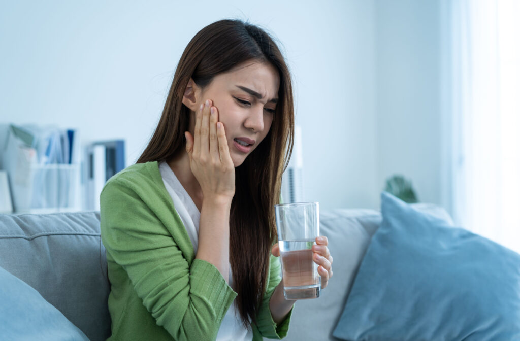 A woman pressing her hand to her cheek because of tooth pain and holding a glass of water.