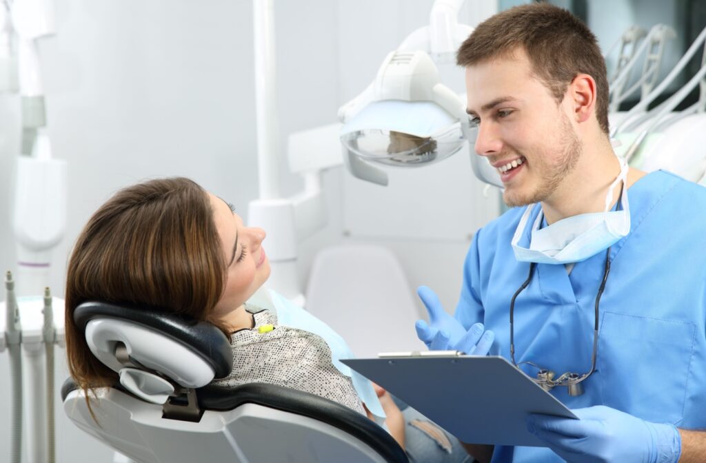A male dentist in blue scrubs holding a clipboard and smiling at a female patient in the dental chair
