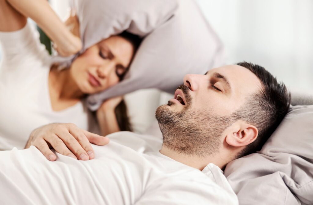 A man snoring in bed while the woman next to him covers her ears with a pillow.