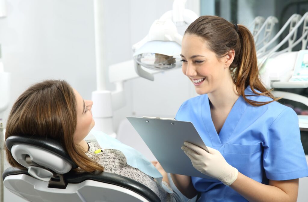 A female dentist in blue scrubs explaining a dental procedure to a woman sitting in a dentist's chair.