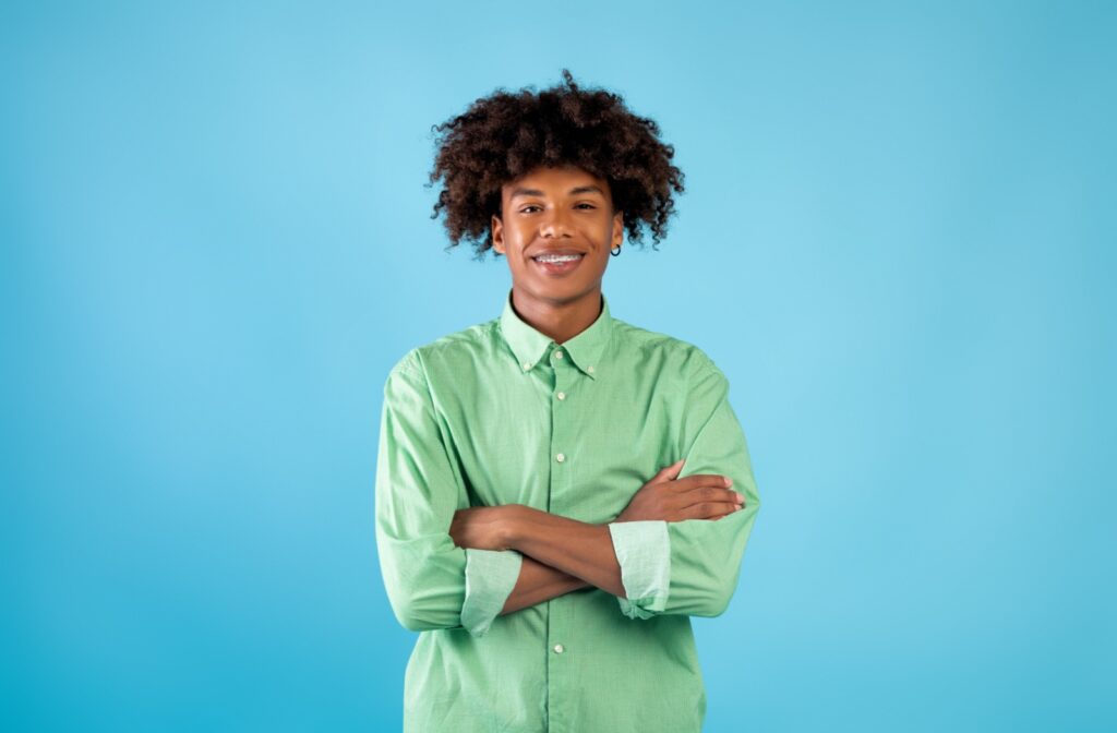 A teenager with braces and a green shirt smiling as he stands with his arms crossed in front of a blue background.