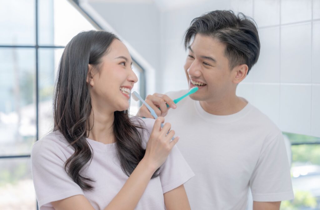 A young couple smiling at each other while they brush their teeth.