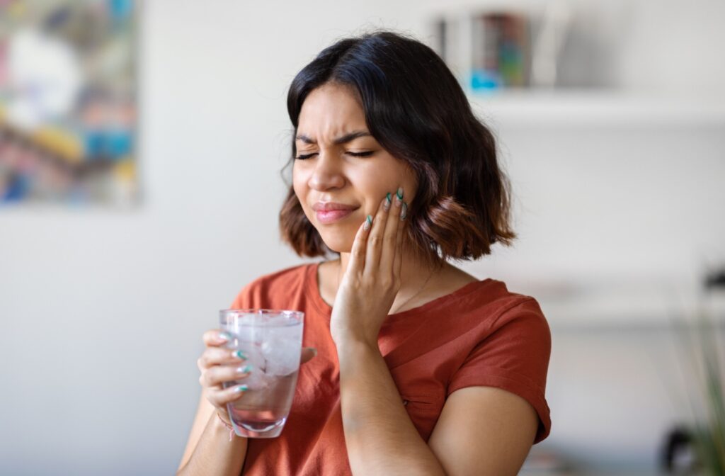 A young woman holding her jaw in pain with her eyes closed while holding a glass of ice water in her right hand.