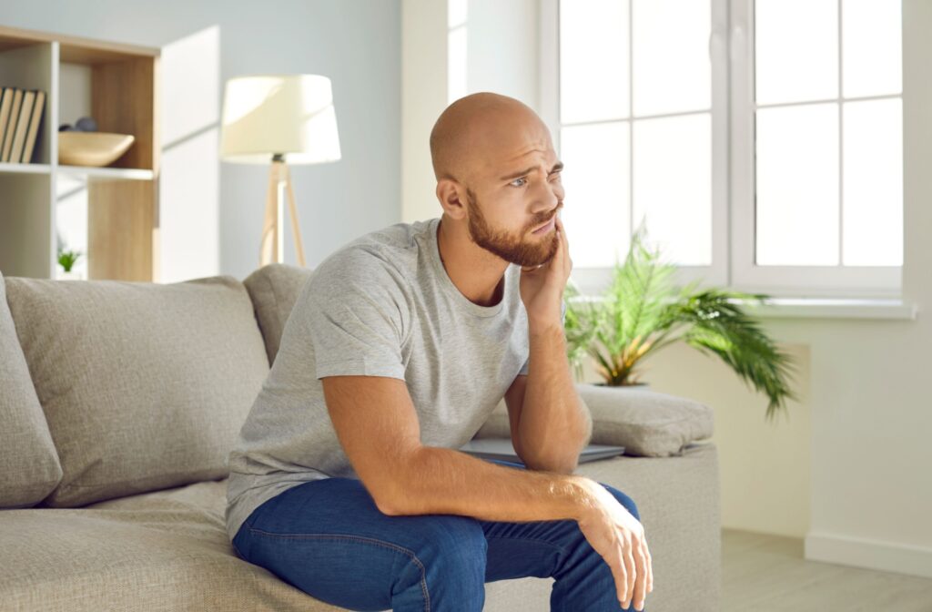 Young person holding cheek and jaw because of tooth pain while sitting on couch at home