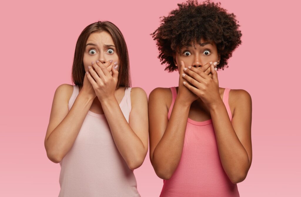 Two adults in shock covering their mouths with their hands in front of a pink background.