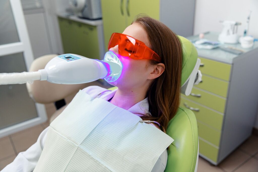 A young person with longer brown hair lays down on a dentist chair, with protective eyewear. They have a special whitening laser in their mouth and are receiving an in-office whitening treatment.