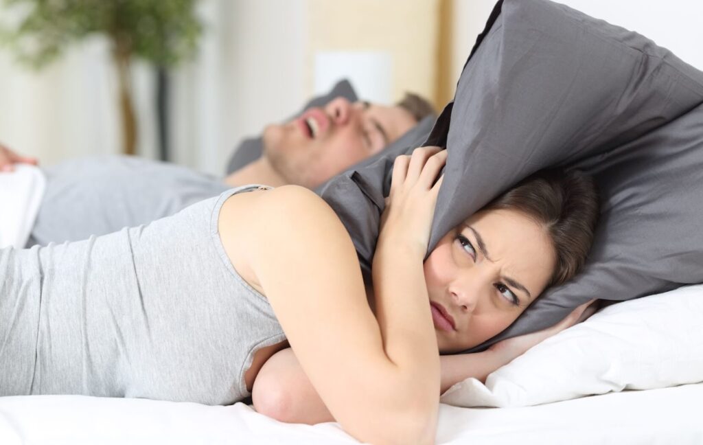 A person with undiagnosed sleep apnea snores loudly as they sleep. Their partner covers their ears while looking irritated.