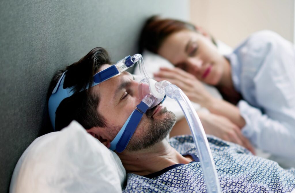 A couple sleeps quietly in their bed. One person is wearing a CPAP machine on their face to help with their sleep apnea.